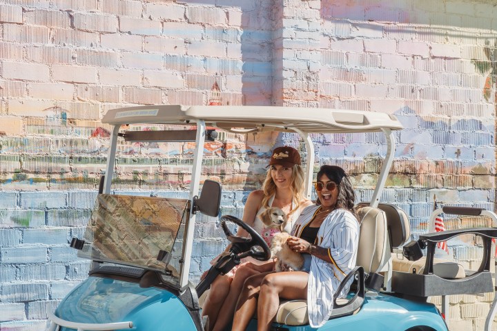 Two people in a turquoise golf cart, parked by a colorful wall with 'Golf Carts' sign.