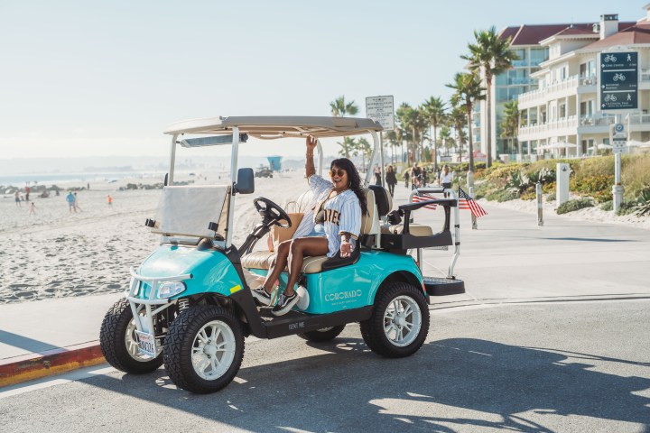 Person waving from turquoise golf cart by beachside road.